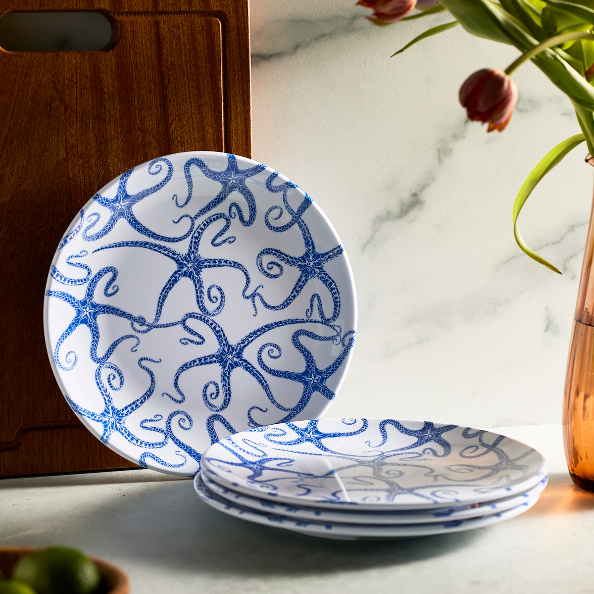 Caskata&#39;s Starfish Melamine Dinner Plates, Set of 4, featuring a blue starfish pattern, are stacked beside a wooden board. Tulips in a vase and limes accent the marble-like surface. Crafted from BPA-free melamine, these plates blend style and safety for your dining experience.