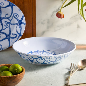 A Starfish Melamine Serving Bowl by Caskata, with its white and blue seaweed pattern, sits elegantly on the counter. Accompanied by a wooden bowl of limes and cutlery, a vibrant red tulip hangs above. Ideal for outdoor entertaining.