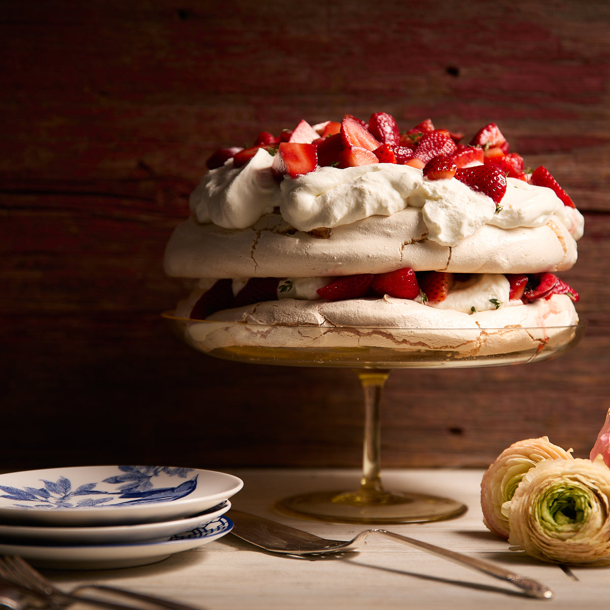 A layered meringue dessert topped with whipped cream and sliced strawberries rests elegantly on a Caskata Celia Citrine Cake Pedestal, next to a stack of plates and flowers on a wooden table.