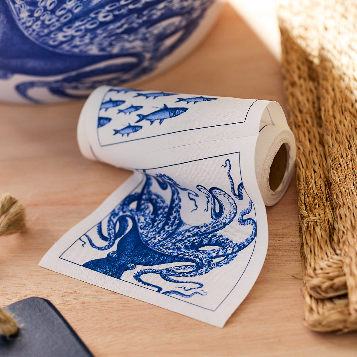 A roll of decorative paper napkins with blue prints of an octopus and fish, placed on a wooden surface next to woven and black items, pairs beautifully with the chic Host With the Most Bundle from Caskata.