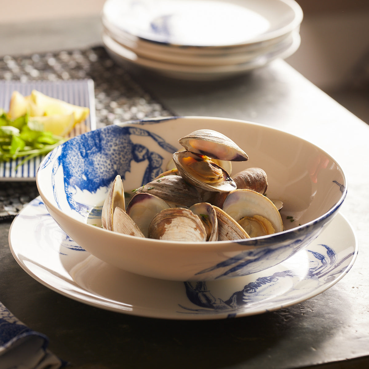 A bowl with a blue and white design is filled with steamed clams, evoking the charm of Caskata&#39;s Lobster Boil Bundle. Plates and green herbs are in the background.
