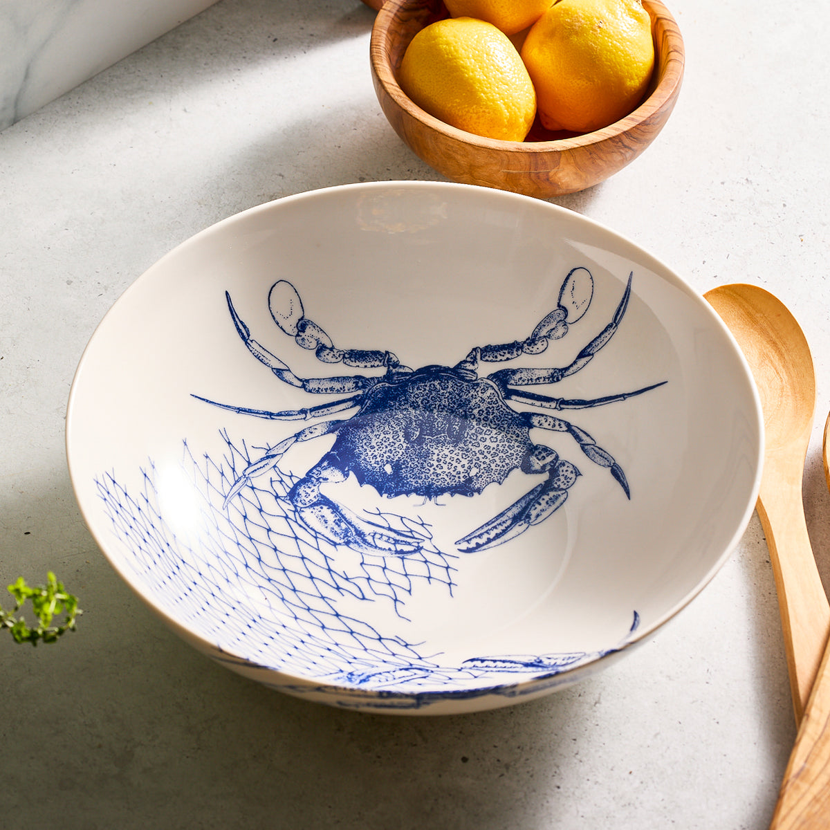 The Crab Wide Serving Bowl by Caskata Artisanal Home features a crab pattern inside, placed on a marble surface with wooden spoons and lemons in the background.