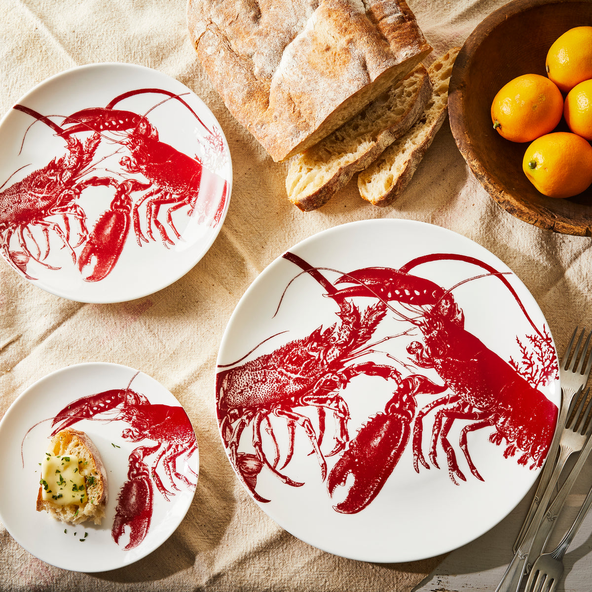 Three white porcelain plates featuring red lobster illustrations from the Beach Wedding Bundle by Caskata are arranged on a table with bread, a wooden bowl of oranges, and a partial view of silverware—a perfect setting for beach lovers.