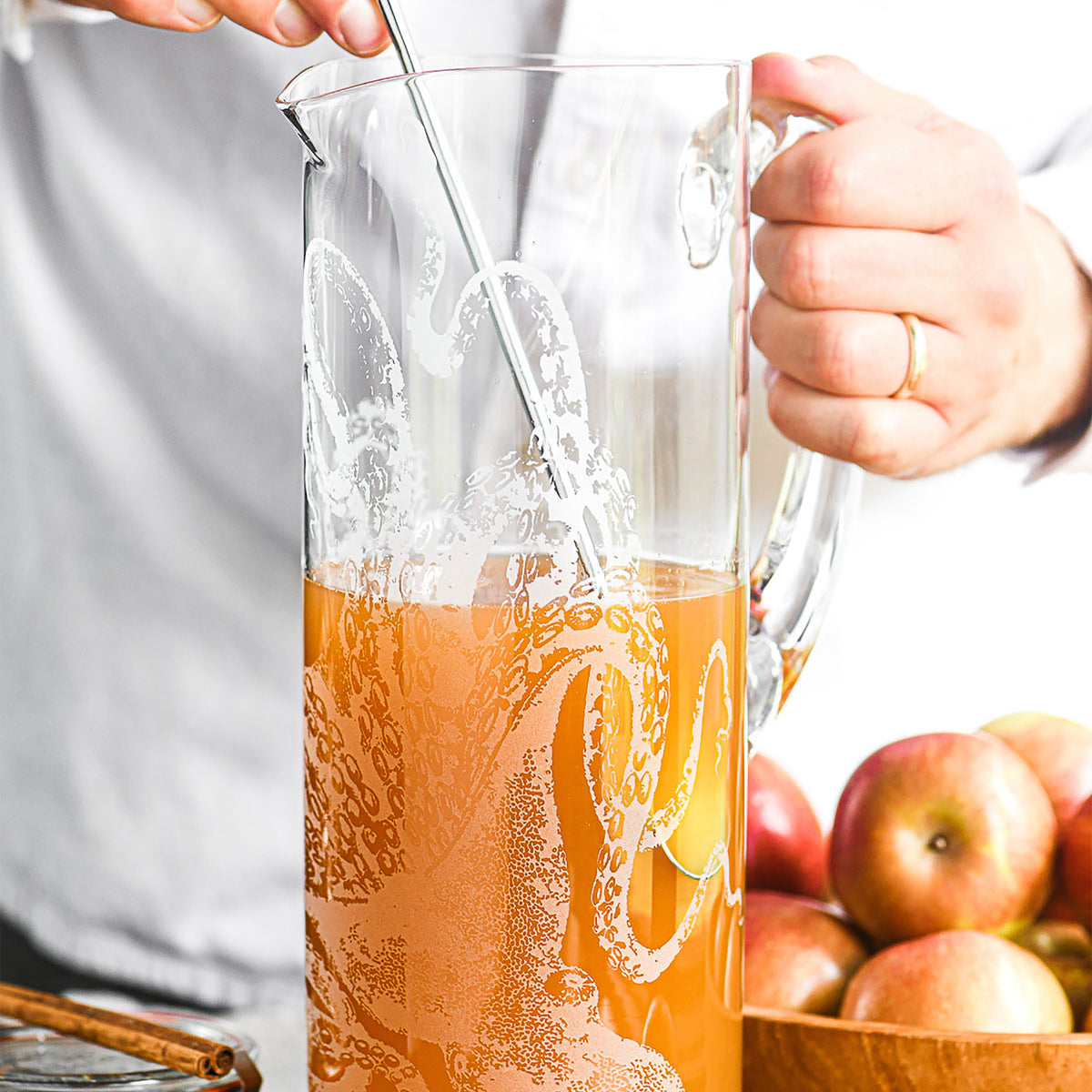 A person stirs apple cider in a Caskata Artisanal Home Lucy Tall Pitcher beside a bowl of apples.