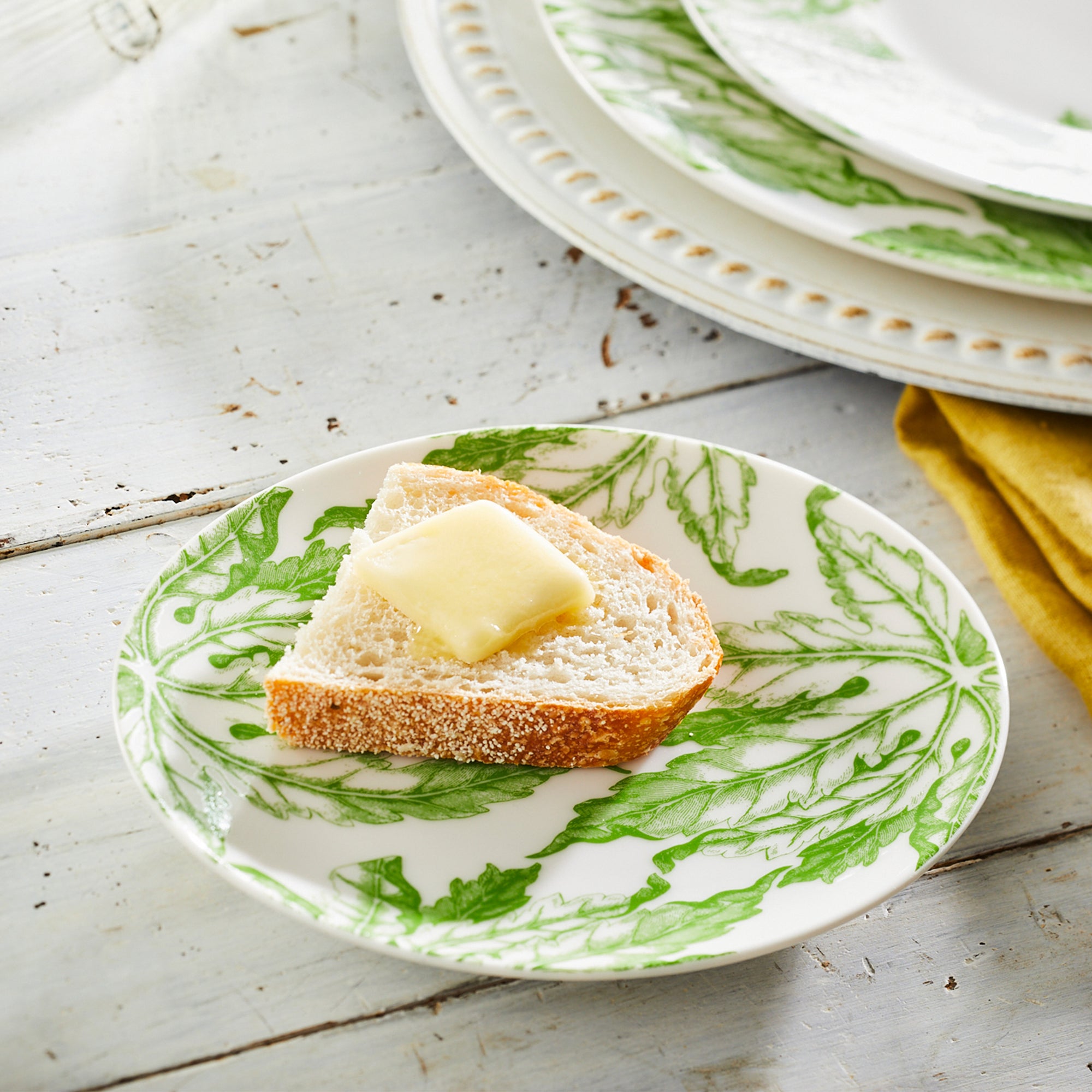 Introducing the Greenhouse Luncheon Bundle by Caskata: Ideal for simple gatherings, it features four white plates with green scalloped edges, four napkins with a charming bird pattern, and floral-design coasters—everything for a spring table refresh.