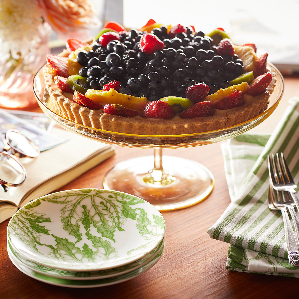 A fruit tart on a Caskata Celia Citrine Cake Pedestal, topped with blueberries and strawberries, surrounded by a book, eyeglasses, a folded green-striped napkin, utensils, and a stack of decorative plates.