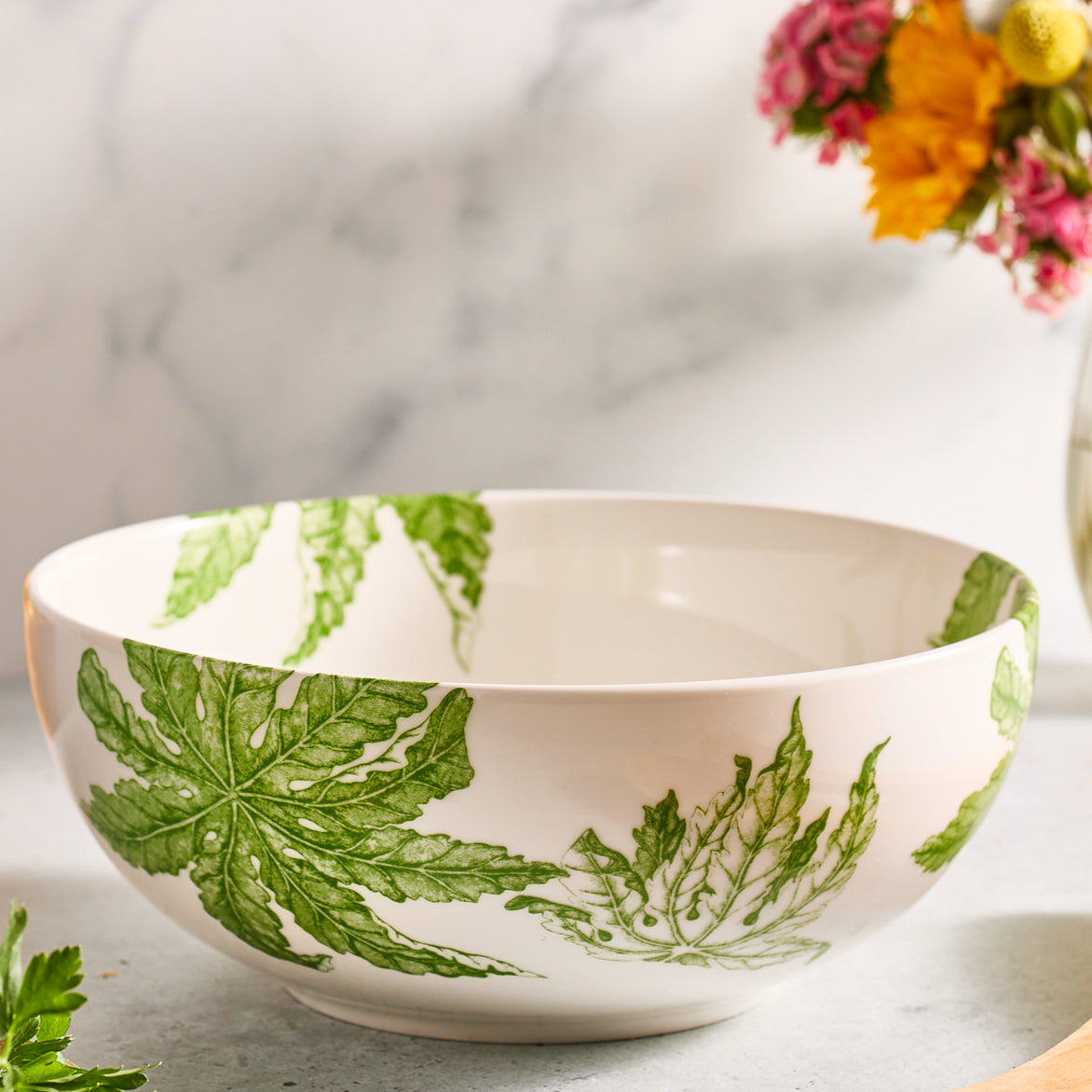 The Freya Vegetable Serving Bowl by Caskata Artisanal Home features delicate green leaf patterns on white ceramic and rests on a light surface. A blurred flower bouquet in the background enhances the bowl&#39;s understated elegance.