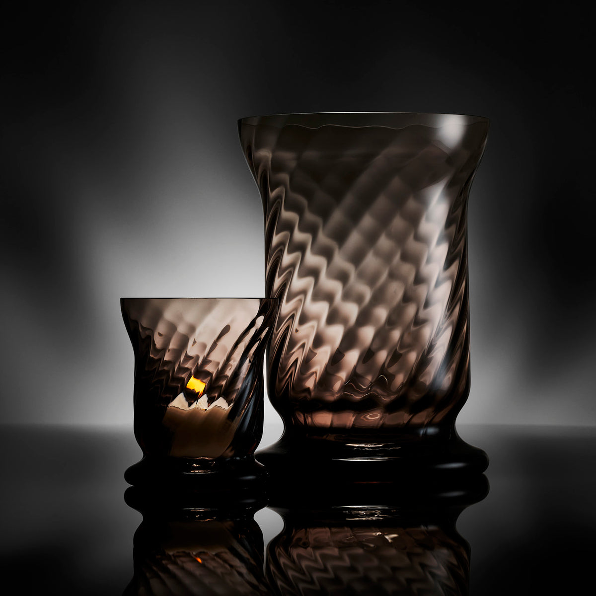 Two textured, amber-tinted vases crafted from mouth-blown glass stand elegantly against a dark backdrop, reflecting light. Their unique sizes and artisanal design evoke the charm of Caskata&#39;s Quinn Mocha Candlelight Bundle, adding sophistication to any setting.