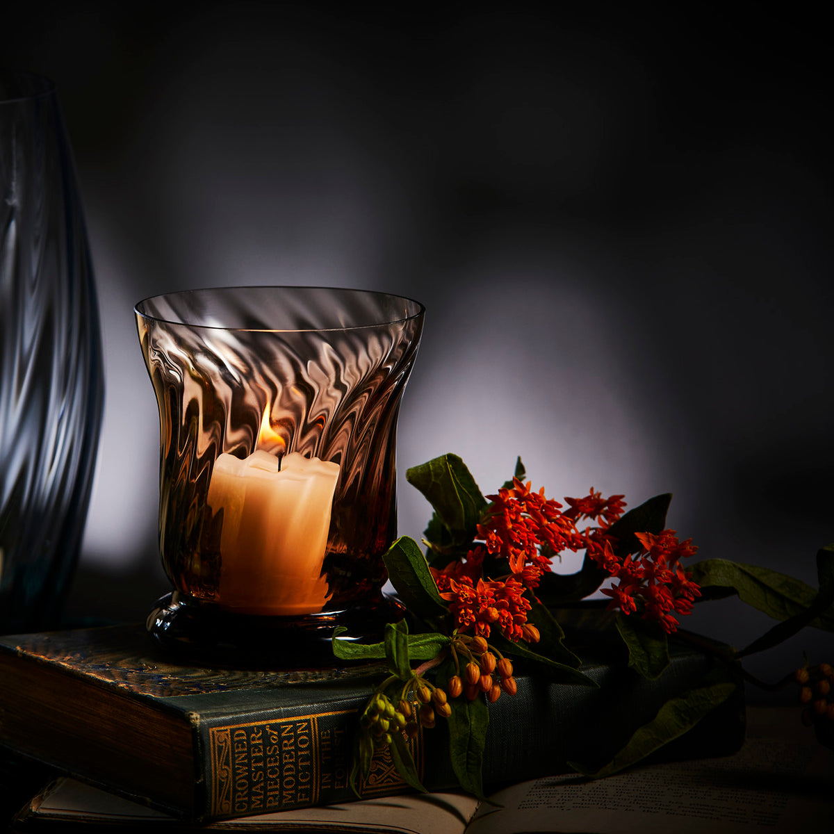 The Quinn Mocha Candlelight Bundle by Caskata features a lit candle in a mouth-blown glass votive on books, beside vibrant red flowers, set against a dimly lit background.