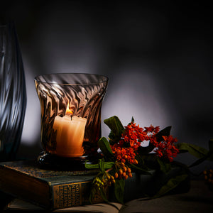 The Quinn Mocha Candlelight Bundle by Caskata features a lit candle in a mouth-blown glass votive on books, beside vibrant red flowers, set against a dimly lit background.