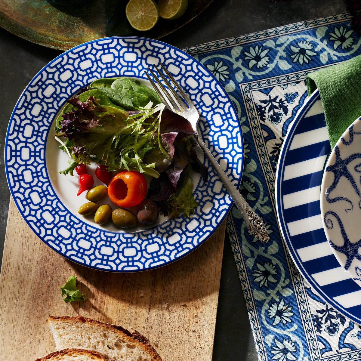 A decorated plate with a fresh salad, including mixed greens, red peppers, and olives, is placed on a wooden board next to a slice of bread and a Caskata Artisanal Home Newport Garden Gate Rimmed Salad Plate on an interlocking lattice pattern tablecloth.