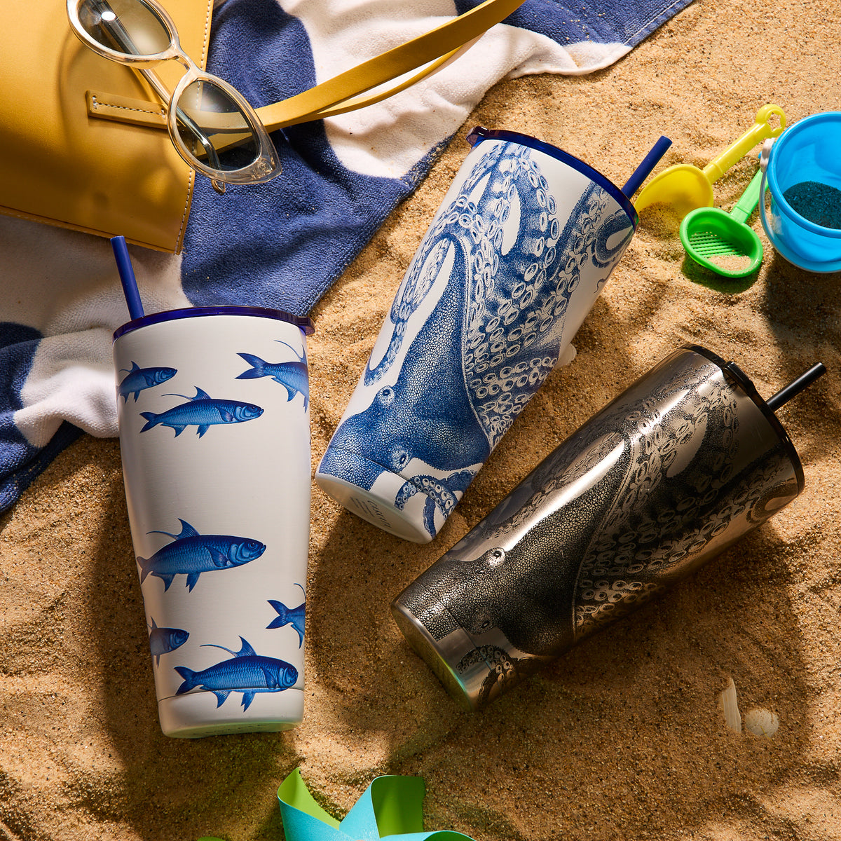 On the sandy beach, three School of Fish Stainless Steel Insulated Tumblers from Caskata with intricate aquatic patterns are accompanied by sunglasses, a towel, and beach toys. Take pleasure in your stylish drinkware collection adventure.