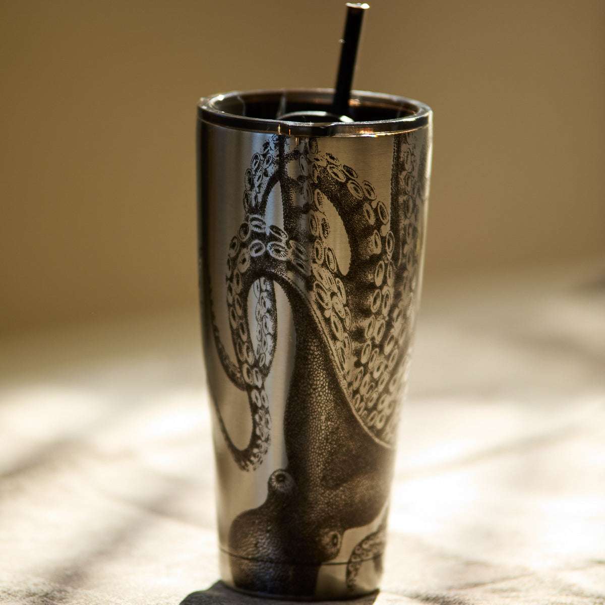 The Lucy Chrome Stainless Steel Insulated Tumbler by Caskata showcases an intricate octopus design and includes a black straw, all set against a blurred background. This vacuum-insulated tumbler maintains your beverages at the ideal temperature while infusing a hint of oceanic elegance.