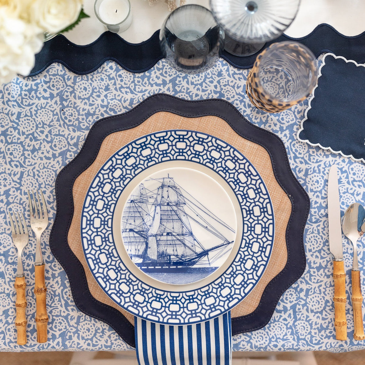 A table setting features a decorative ship-themed blue and white plate surrounded by utensils with bamboo-like handles on a patterned cloth. The Caskata Pinstripe Dinner Napkins in Blue Set/4 adds charm, complementing the blue theme while ensuring elegance with their 100% cotton fabric.