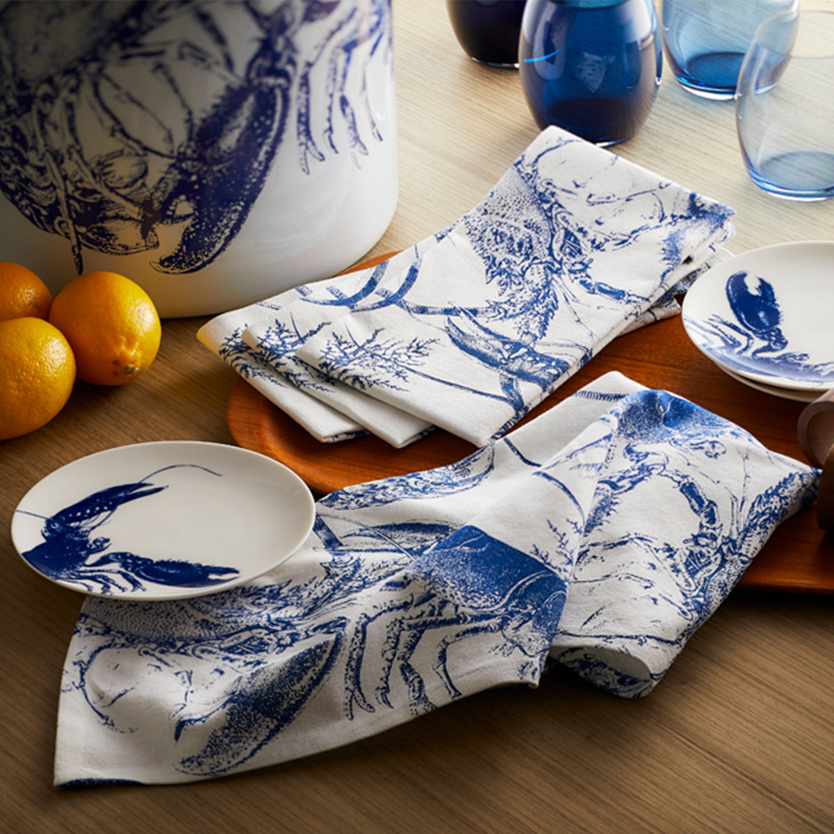 A table setting with crab-themed napkins, Blue Lobster Small Plates, blue glasses, Les Nuages Tumblers, a large ceramic container, and a wooden tray holding lemons from the Host With the Most Bundle by Caskata.