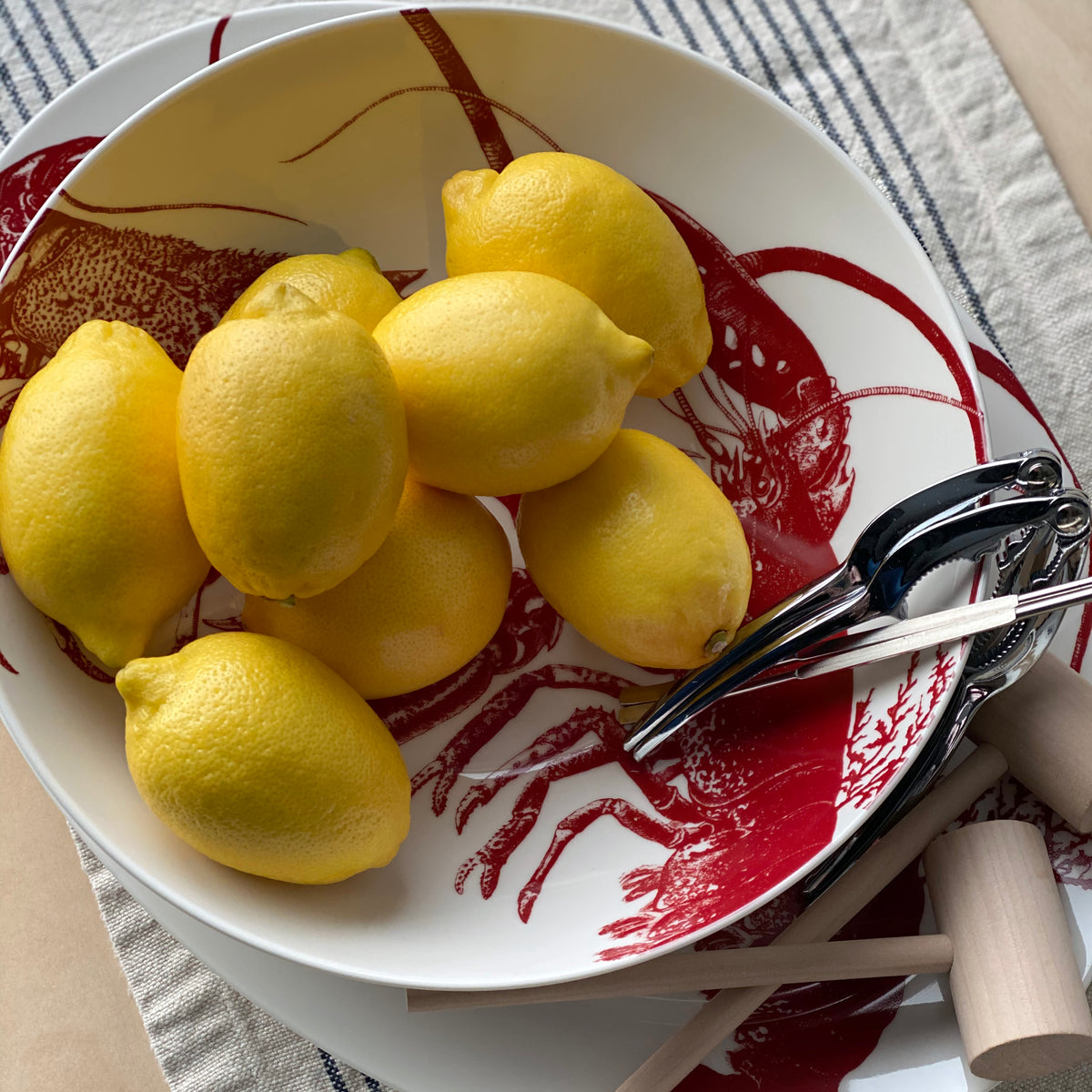 A white plate with a red lobster print holds several yellow lemons and a metal citrus juicer. The plate is placed on a striped tablecloth, perfectly complementing the July Fourth Bundle by Caskata nearby.