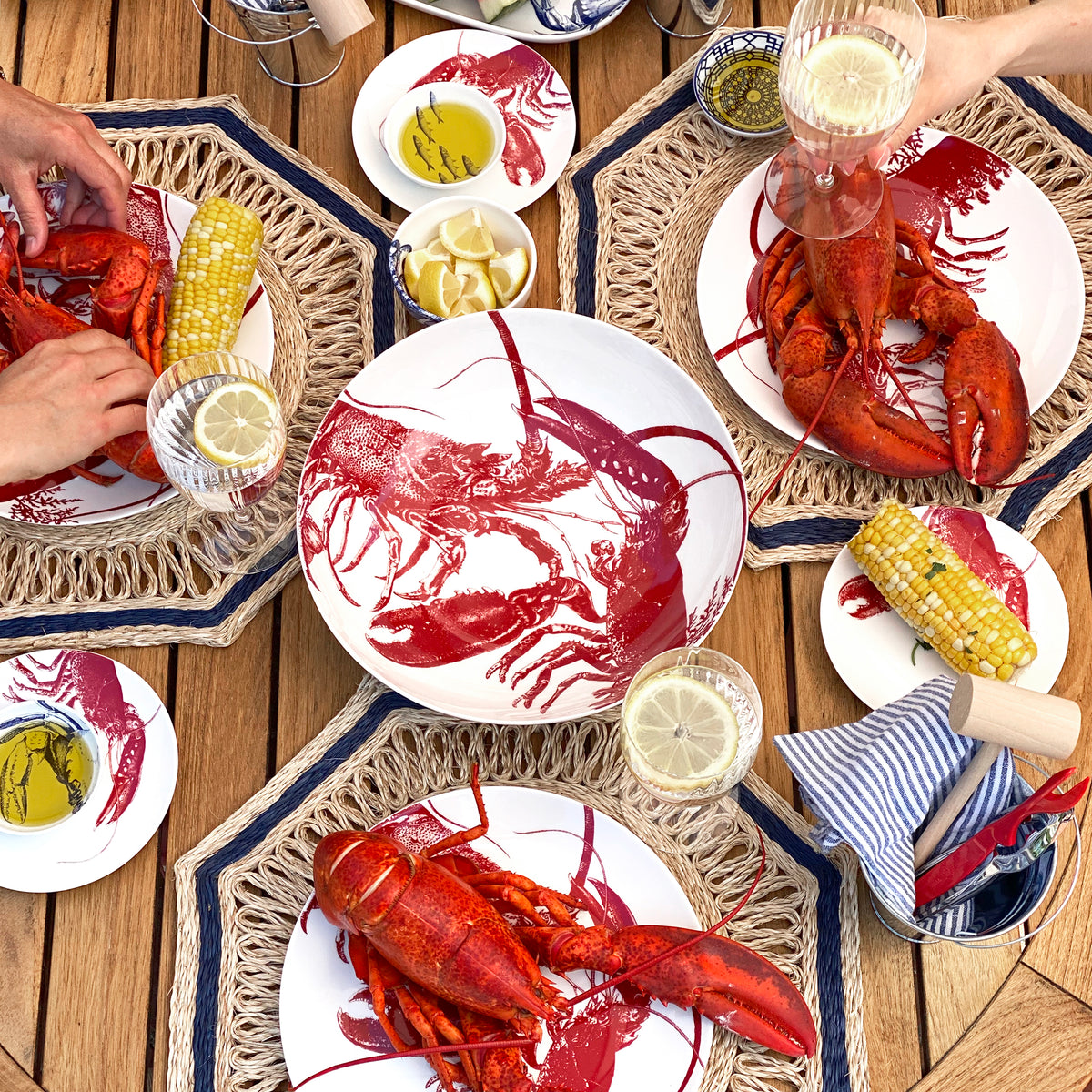 Four place settings with lobster-themed dishes and placemats feature whole lobsters, corn on the cob, and lemon wedges on a wooden table. A July Fourth Bundle by Caskata brimming with fresh seafood sits in the center. Two hands are visible, holding a lobster and a glass of water.