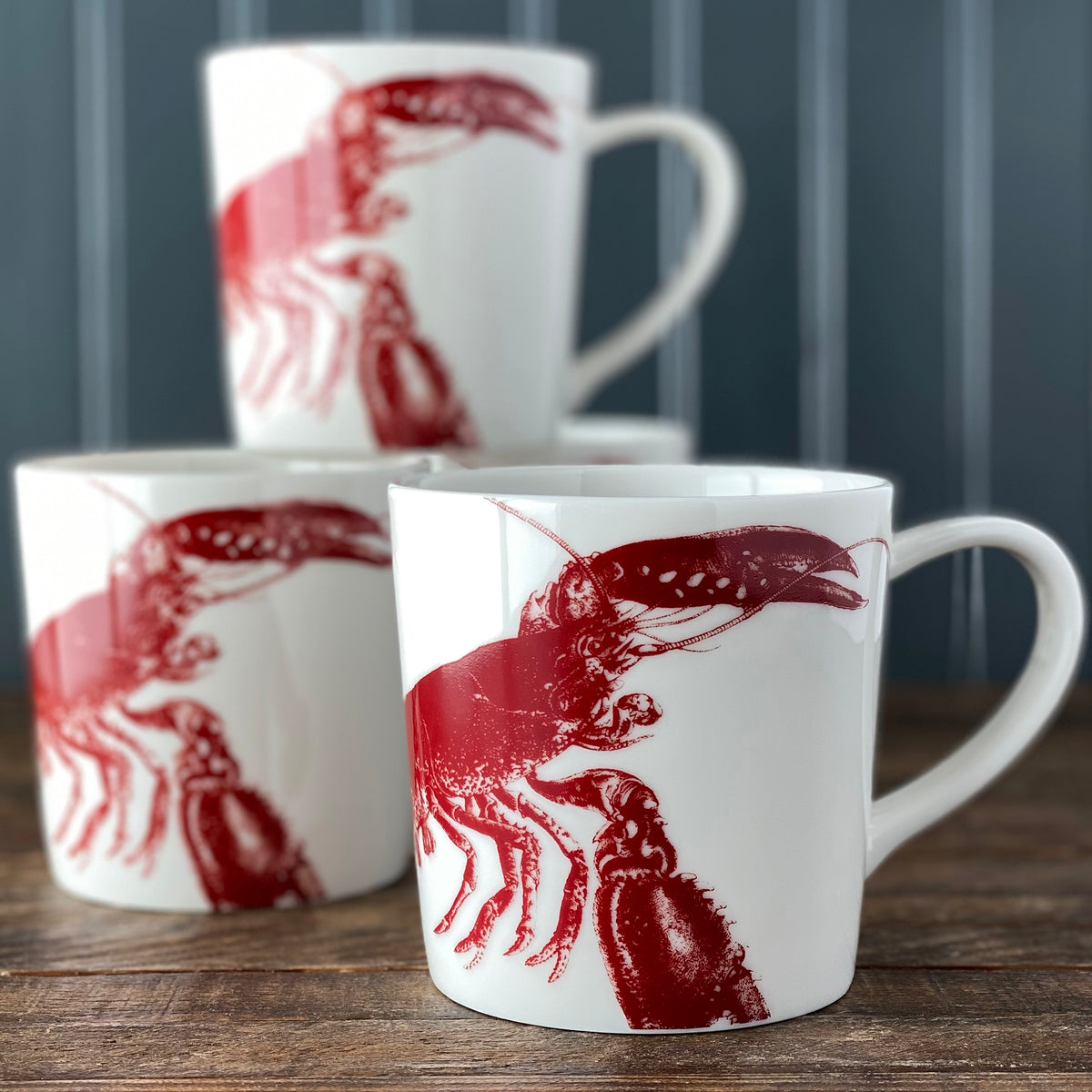 Three white ceramic &quot;Father&#39;s Day Bundle&quot; mugs with red designs from Caskata are stacked on a wooden surface against a dark vertical striped background.