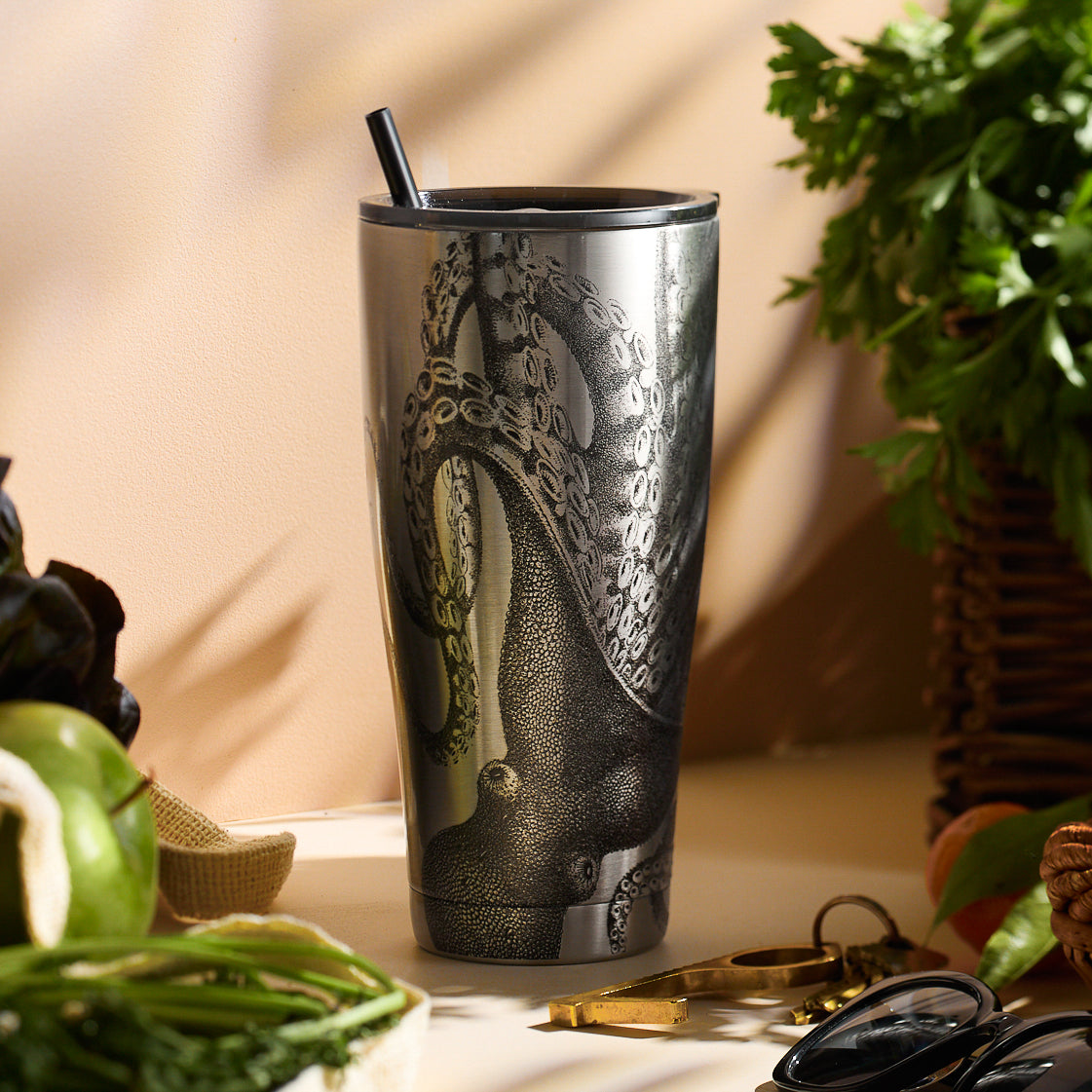 The Lucy Chrome Stainless Steel Insulated Tumbler by Caskata, featuring an octopus design, stands on a counter surrounded by vegetables and eyeglasses.