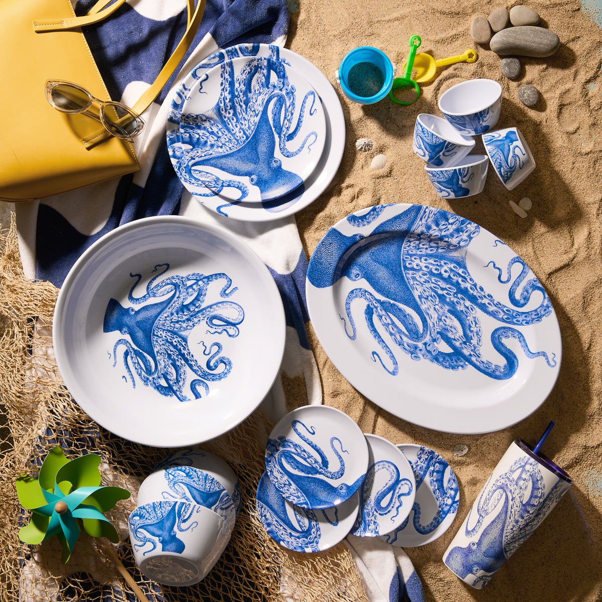 The Lucy Melamine Oval Rimmed Platter by Caskata, featuring a blue and white octopus theme, is arranged on sand with a yellow bag, sunglasses, pinwheel, and beach toys. This BPA-free dinnerware set is perfect for seaside picnics and outdoor use.