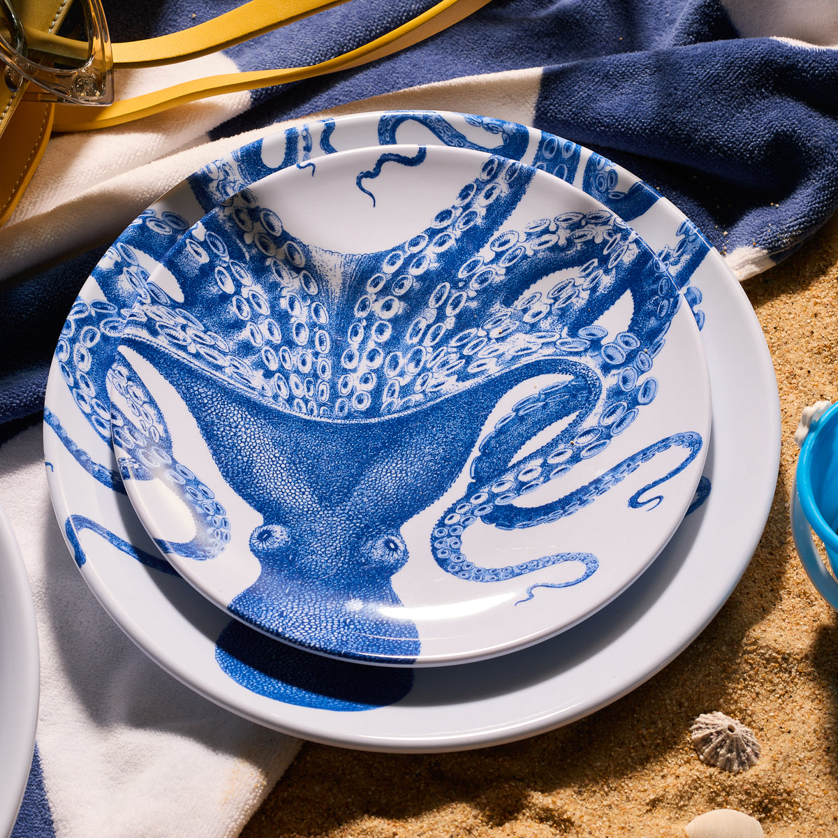 Two Lucy Melamine Salad Plates from Caskata, featuring a charming blue octopus design, are stacked on a towel nestled on the sand with seashells nearby. Made from BPA-free melamine, these unbreakable plates ensure worry-free beachside dining.