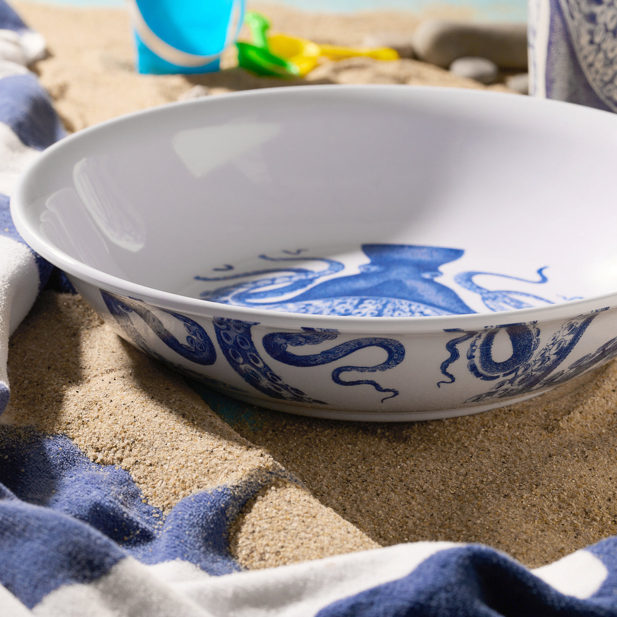 The Lucy Melamine Serving Bowl by Caskata, with a blue octopus design, rests on sandy beach towels amidst beach toys—part of our octopus dinnerware collection.