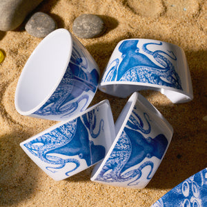 A set of four Caskata Lucy Melamine Snack Bowls with blue octopus designs rests gracefully on the sand, surrounded by small stones.