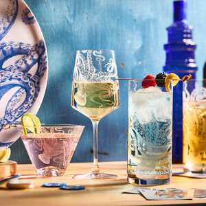 Three Lucy Stemless Martini Glasses from Caskata Artisanal Home grace a wooden table. One shimmers with wine, another holds a cocktail with berries, and the third displays a mixed drink topped with lime slices, alongside a blue octopus-patterned plate.