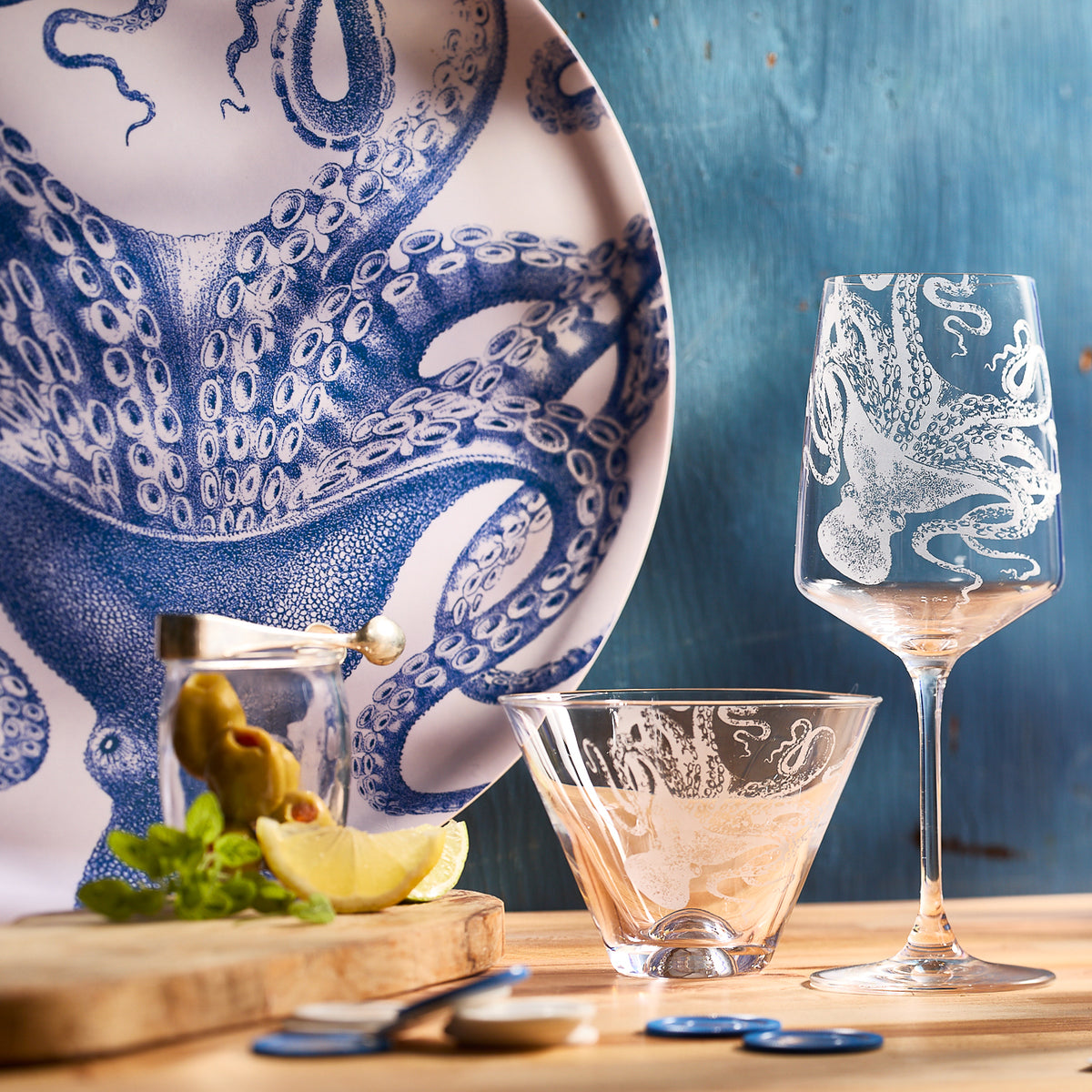 Caskata Artisanal Home&#39;s Lucy Stemless Martini Glasses feature intricate blue designs crafted from lead-free crystal. A wooden board holds olives, lemon, and mint against a textured blue background to enhance the oceanic theme.