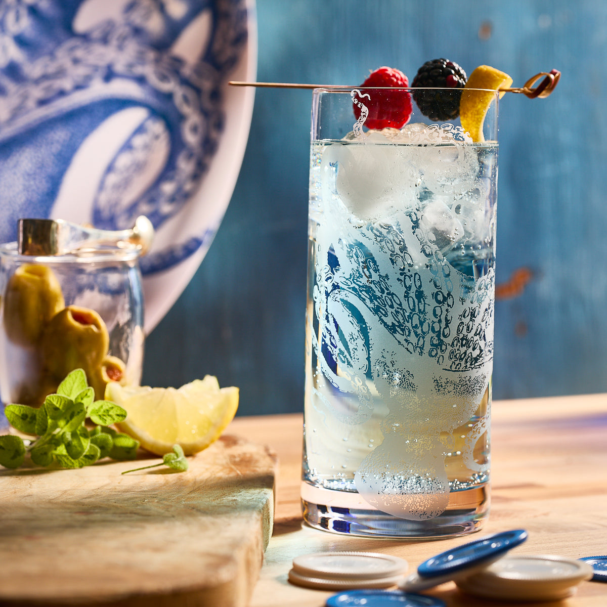 A Caskata Artisanal Home Lucy Highball glass, perfected with sand-etching, is filled with a fizzy drink and ice. Garnished with a lemon twist and berries on a skewer, it sits beside a pitcher, lemon wedge, and mint leaves on a wooden surface.