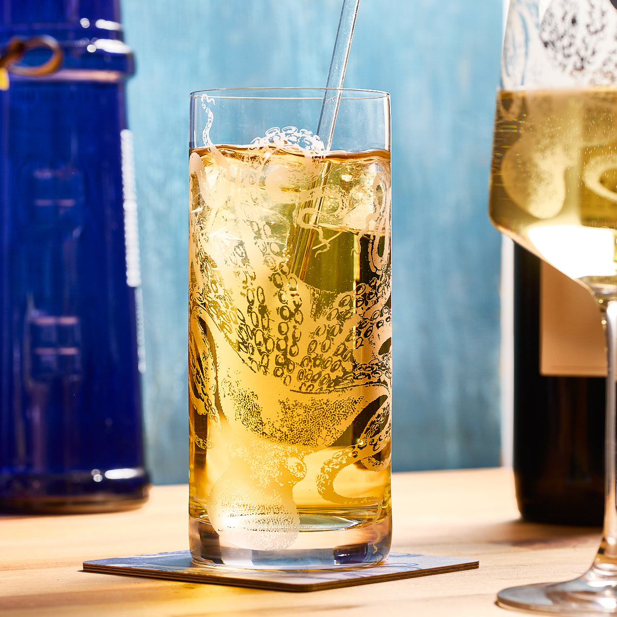 A Lucy Highball Glass from Caskata Artisanal Home, filled with iced drink and showcasing a sand-etched octopus design, is on a coaster. A champagne flute and blue bottle sit nearby on the wooden surface against a light blue backdrop.