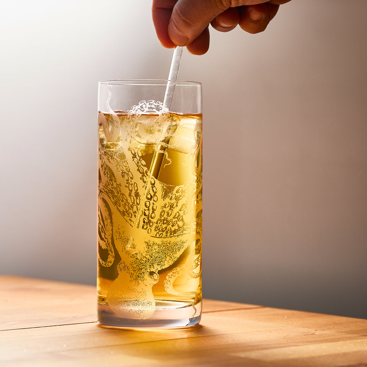 A hand stirs a light brown drink in a Lucy Highball Glass by Caskata Artisanal Home, featuring an octopus design, on a wooden table.