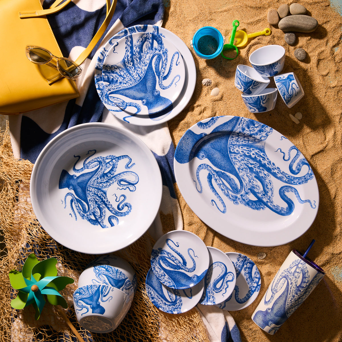 The Lucy &quot;Party Pack&quot; Melamine Bundle by Caskata, a BPA-free and unbreakable blue and white octopus dinnerware set, is artfully arranged on sand. It includes plates, bowls, a tumbler, a yellow bag with sunglasses, and a small beach toy set.