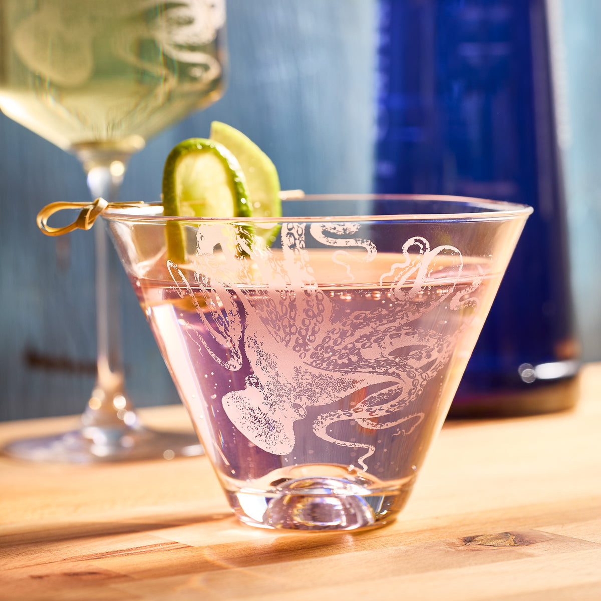 A pink cocktail in Lucy Stemless Martini Glasses by Caskata Artisanal Home, adorned with cucumber slices on a stick, is on a wooden table alongside a blue bottle and another drink. The lead-free crystal&#39;s clarity enhances the visual allure of this martini presentation.