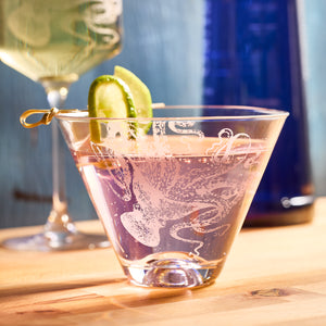 A pink cocktail in Lucy Stemless Martini Glasses by Caskata Artisanal Home, adorned with cucumber slices on a stick, is on a wooden table alongside a blue bottle and another drink. The lead-free crystal's clarity enhances the visual allure of this martini presentation.