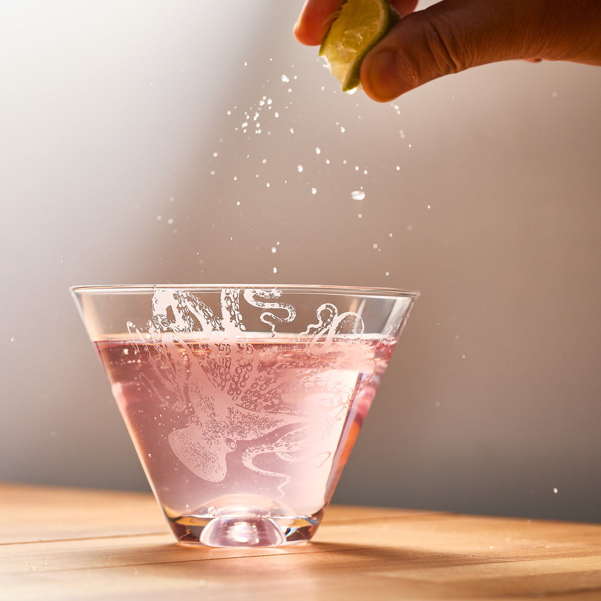 A hand squeezes a lime over a pink liquid in a Lucy Stemless Martini Glass by Caskata Artisanal Home, causing droplets to splash. The lead-free crystal glass rests elegantly on wood against a neutral backdrop.