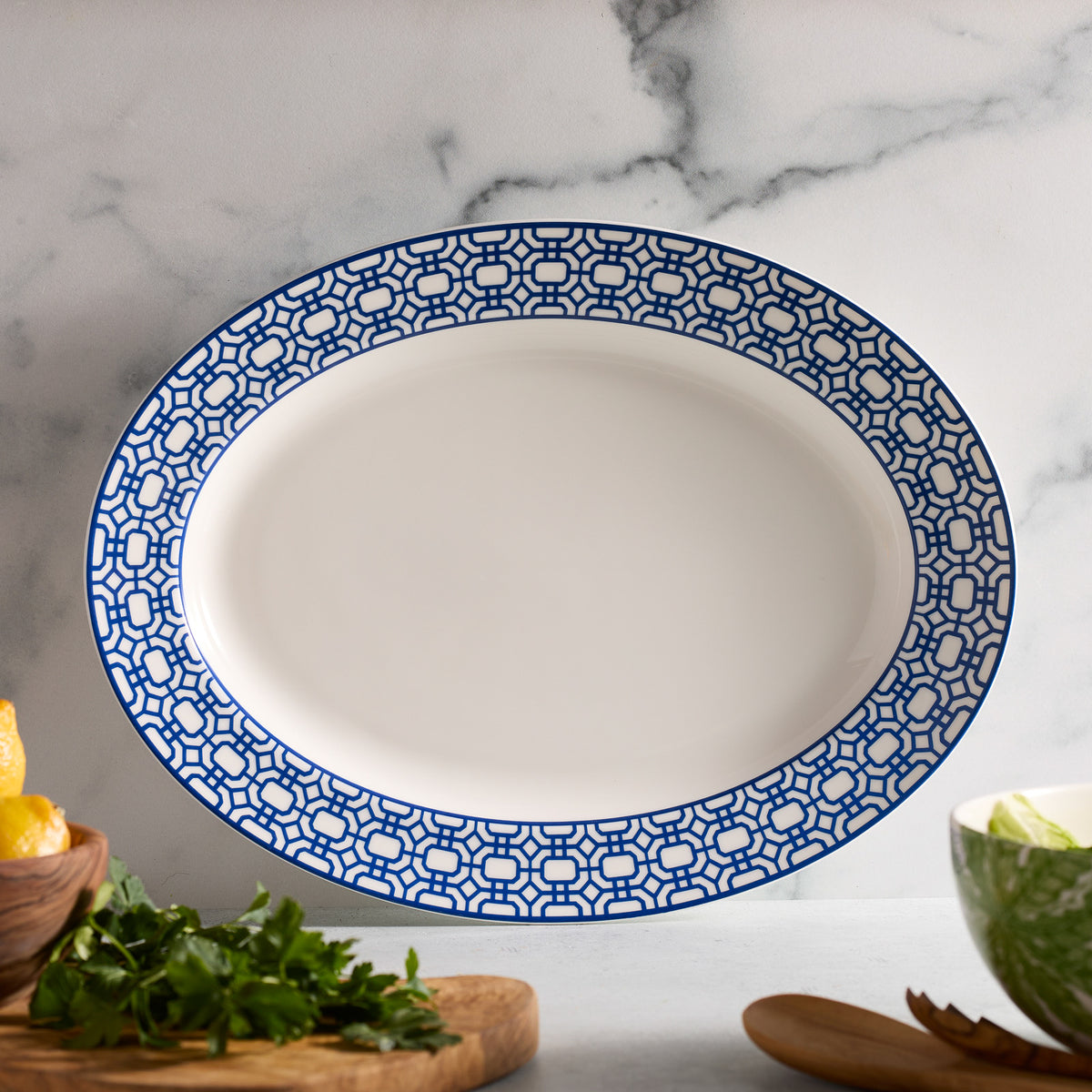 The Caskata Artisanal Home Newport Garden Gate Oval Rimmed Platter, with a blue geometric border on white porcelain, sits on marble. Herbs, a lemon, cabbage, and a wooden spoon create an elegant scene reminiscent of classic designs.