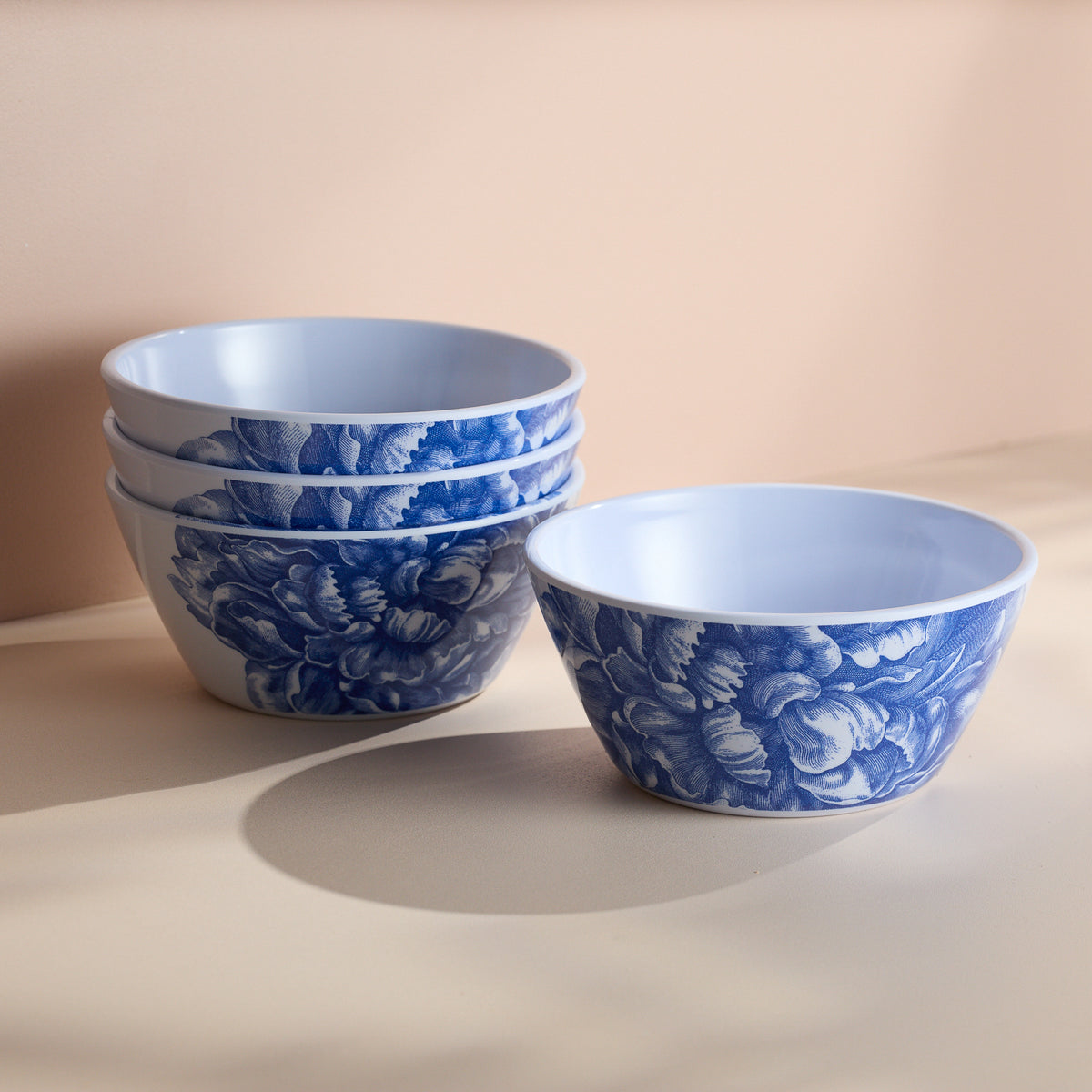 The Caskata Peony Melamine Cereal Bowls, a BPA-free set of four with blue floral patterns, are elegantly stacked on a beige surface, ideal for indoor and outdoor use.