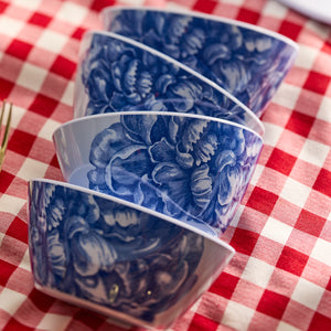 The Peony Melamine Cereal Bowls by Caskata, a set of four with a blue floral pattern and BPA-free, are stacked on a red and white checkered tablecloth. This charming dinnerware set is perfect for both indoor and outdoor use.