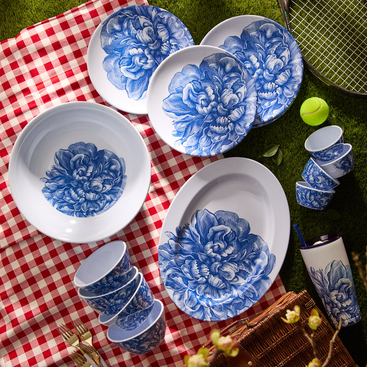 A Peony Melamine Oval Rimmed Platter by Caskata graces a red and white checkered picnic blanket, with a tennis racket and ball nearby, adding an elegant touch to the outdoor scene.