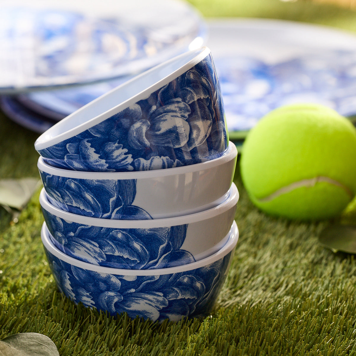 The Peony Melamine Snack Bowls, a set of four by Caskata, feature a blue floral design and are elegantly stacked on artificial grass next to a tennis ball. Made from BPA-free melamine, this unbreakable dinnerware ensures durability and style for any occasion.