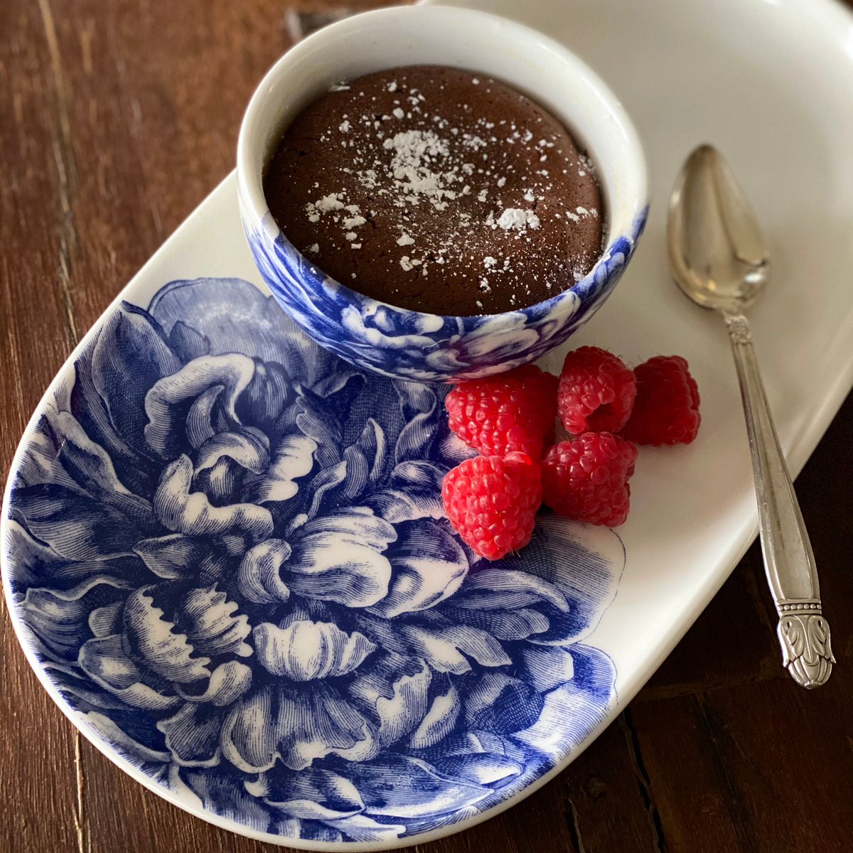 A chocolate dessert in a blue and white floral-patterned bowl on a Peony Small Oval Tray by Caskata Artisanal Home, accompanied by a spoon and four raspberries, all pieces from our exquisite blue and white dinnerware collection.