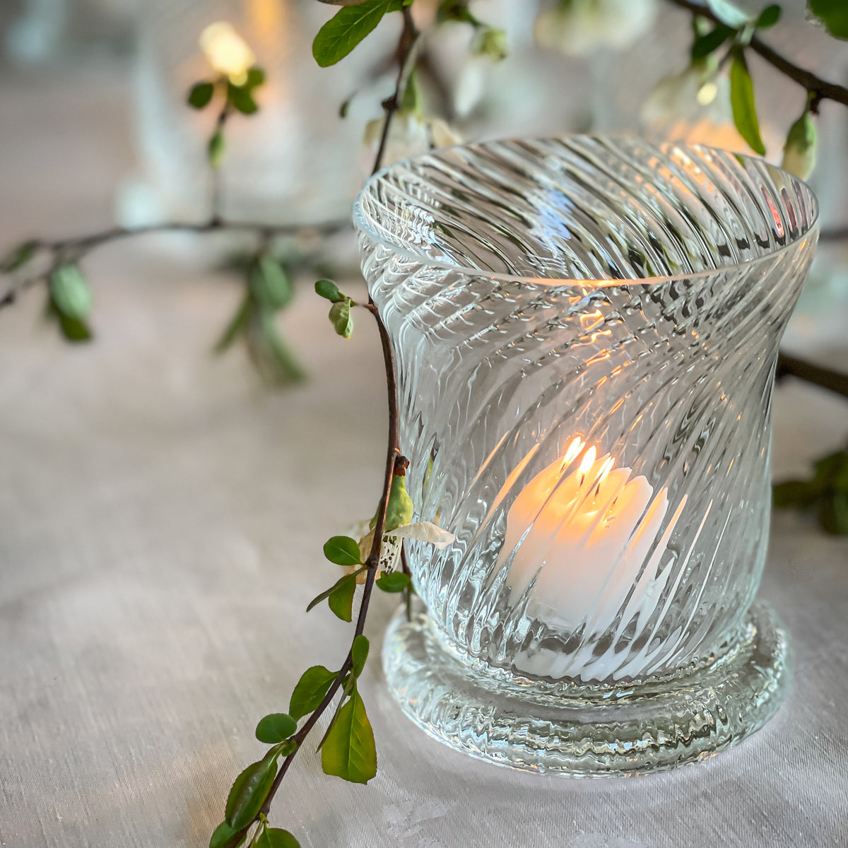 A lit candle from the Caskata Quinn Clear Candlelight Bundle sits on a table next to green branches with small leaves and white flowers.