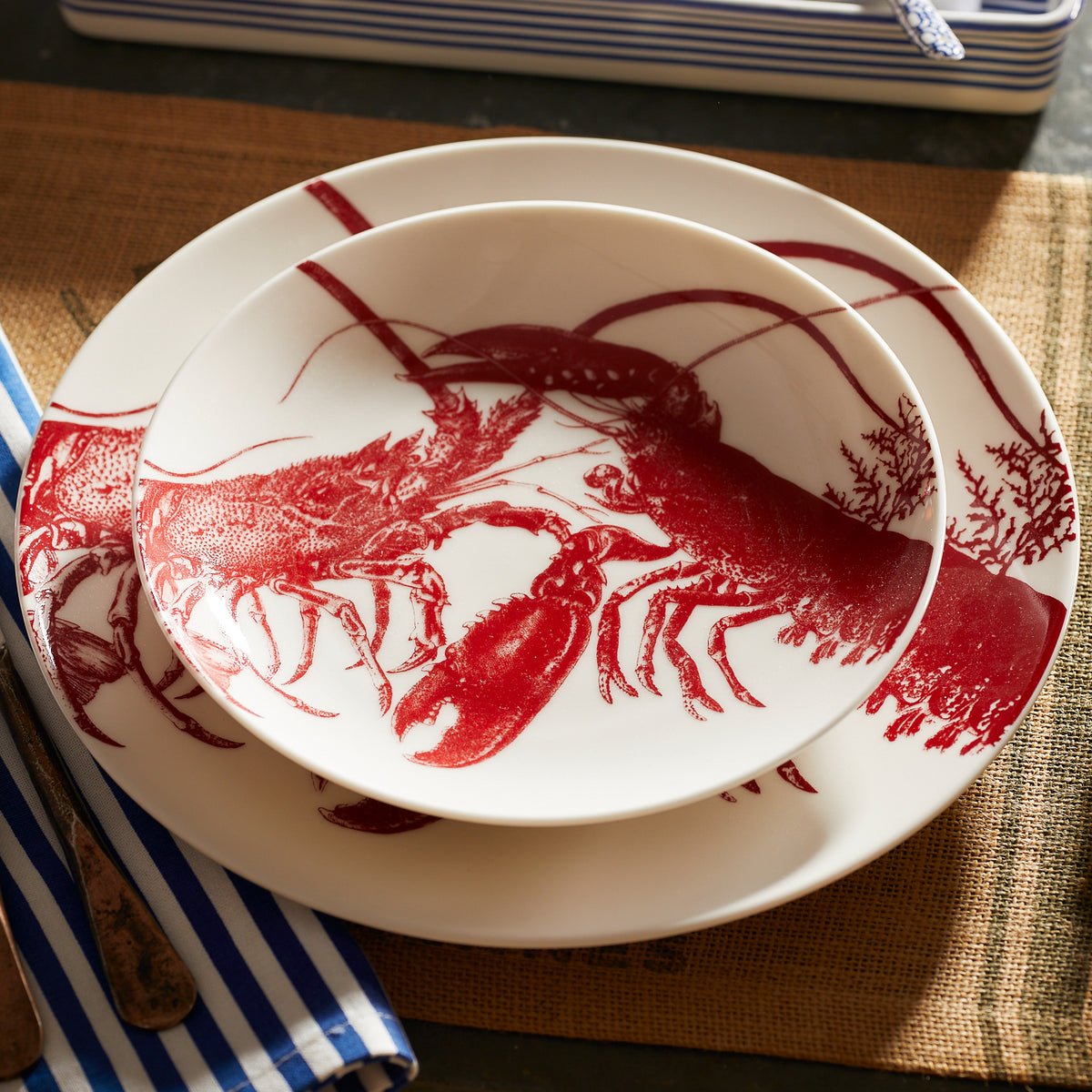 A round dinner plate and a matching shallow bowl, both featuring red lobster illustrations, are set on a woven placemat with a blue and white striped napkin beside them. This stunning Beach Wedding Bundle by Caskata is made from lead-free porcelain and is both dishwasher and microwave safe.