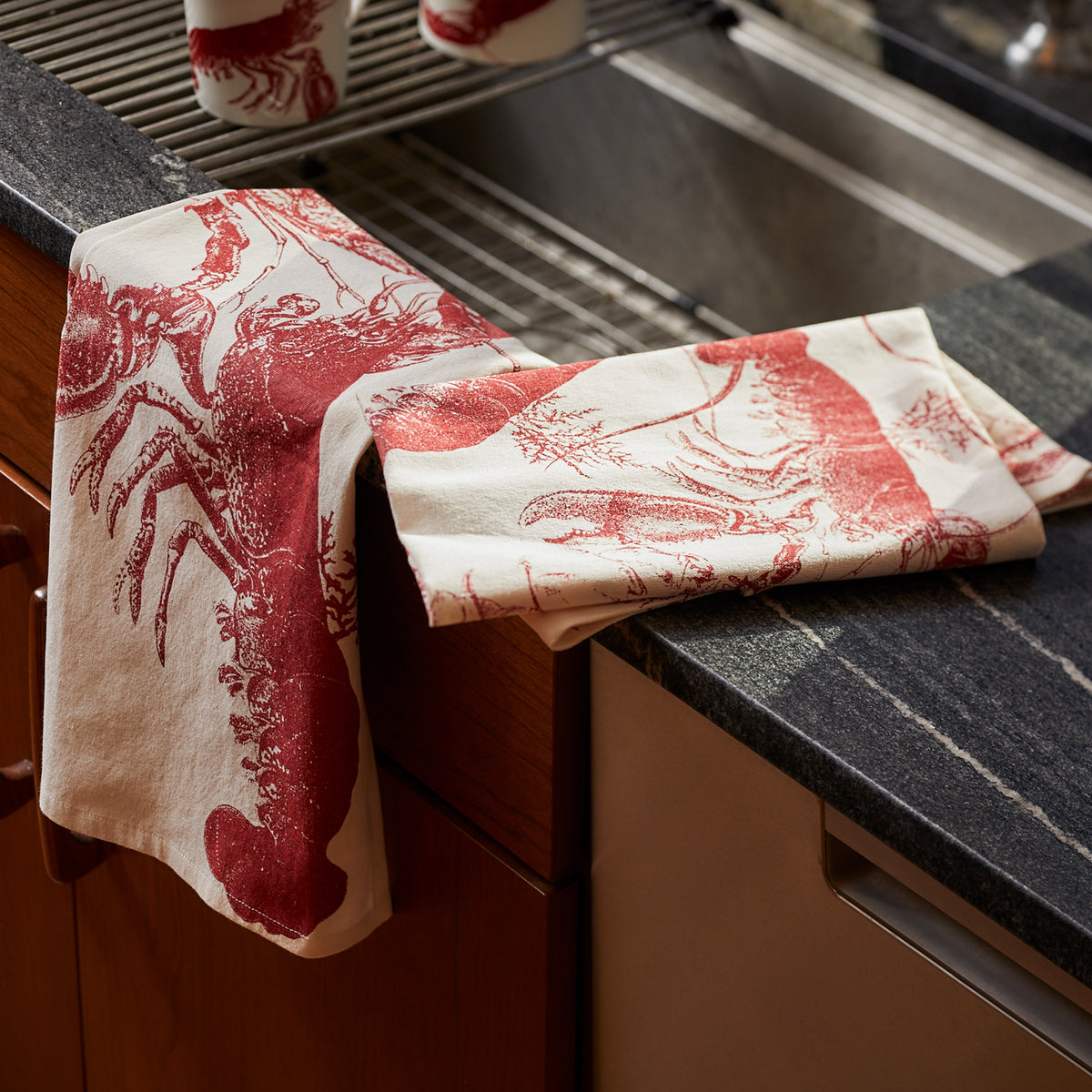 Two white July Fourth Bundle Kitchen Towels by Caskata, featuring red lobster prints, are draped over the edge of a kitchen sink and countertop.