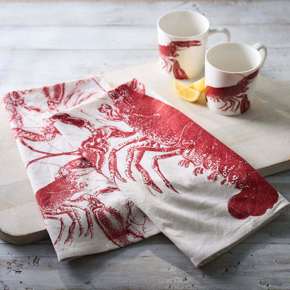 Two Caskata Father&#39;s Day Bundle mugs with red prints sit next to a lemon wedge and a matching lobster kitchen towel on a white wooden surface.