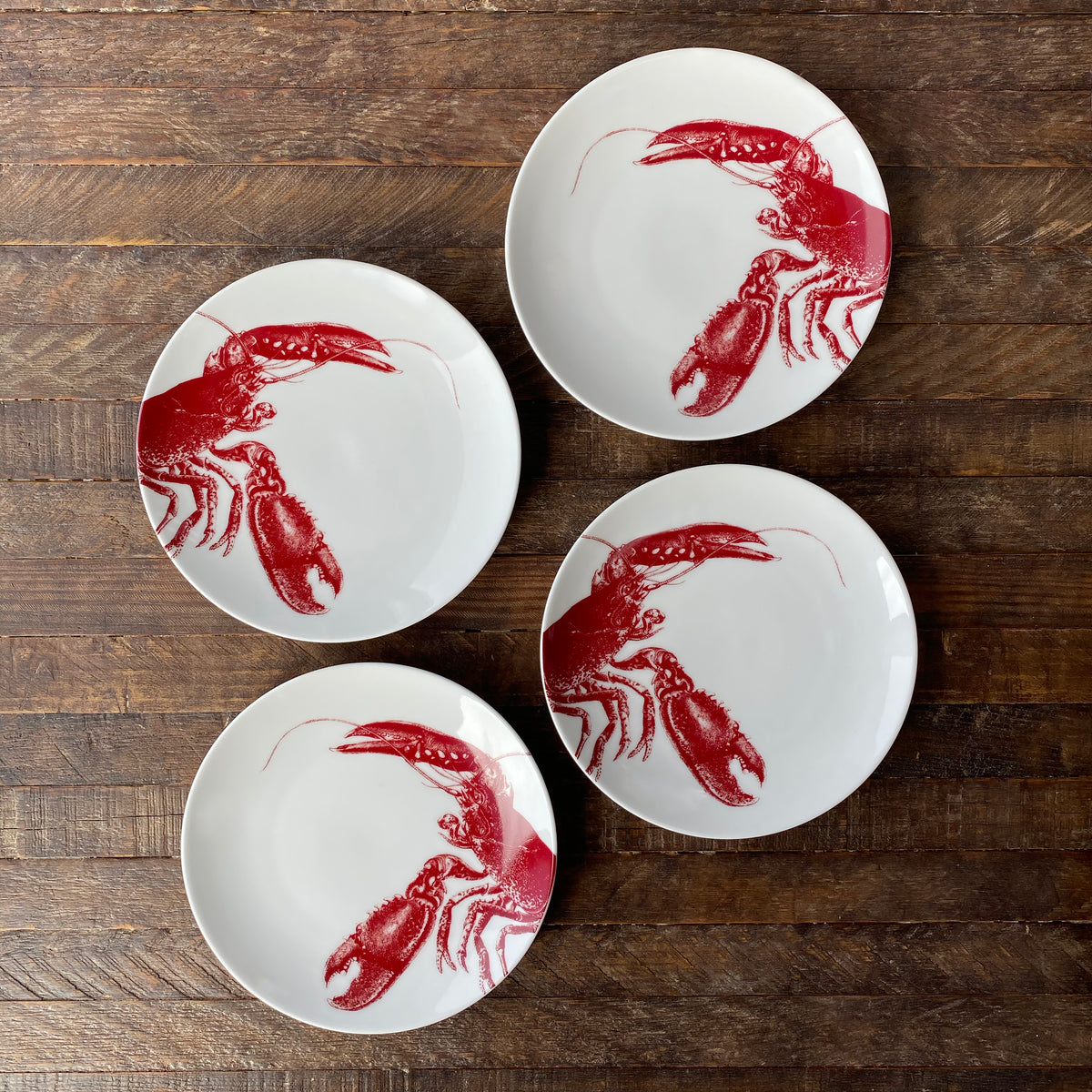 Four porcelain plates, each showcasing a red lobster illustration, are arranged in a circle on a wooden surface. This elegant Beach Wedding Bundle by Caskata adds a touch of coastal charm to your table setting.
