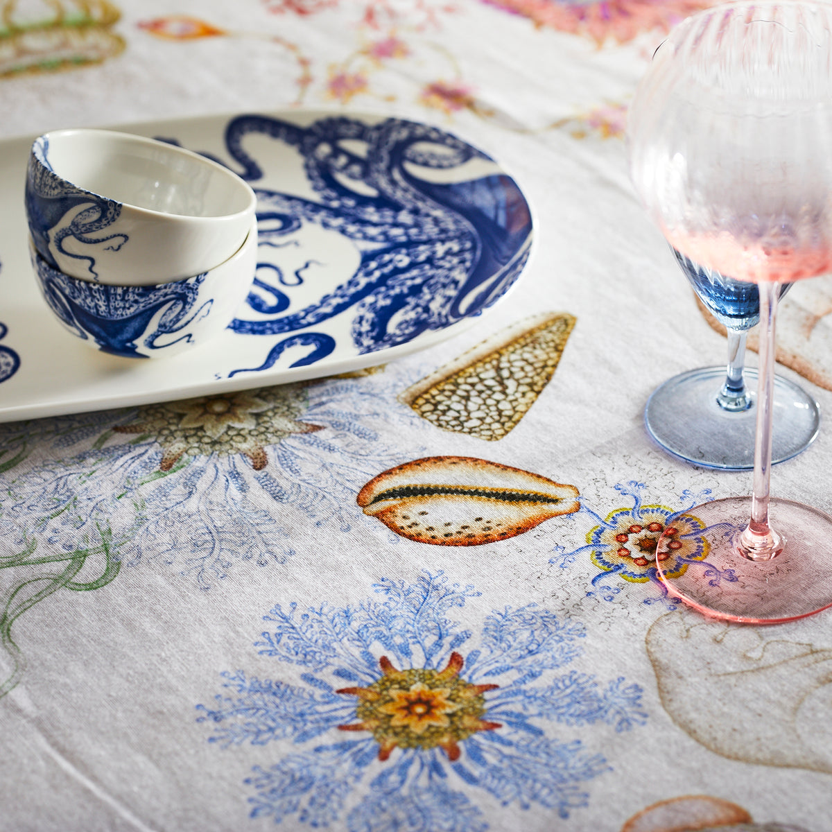 A table set with a detailed sea-themed white tablecloth, a white cup and saucer made of premium porcelain featuring blue sea creatures, two elegant stemmed glasses, one pink and one blue, and a Lucy Snack Bowl from Caskata Artisanal Home.