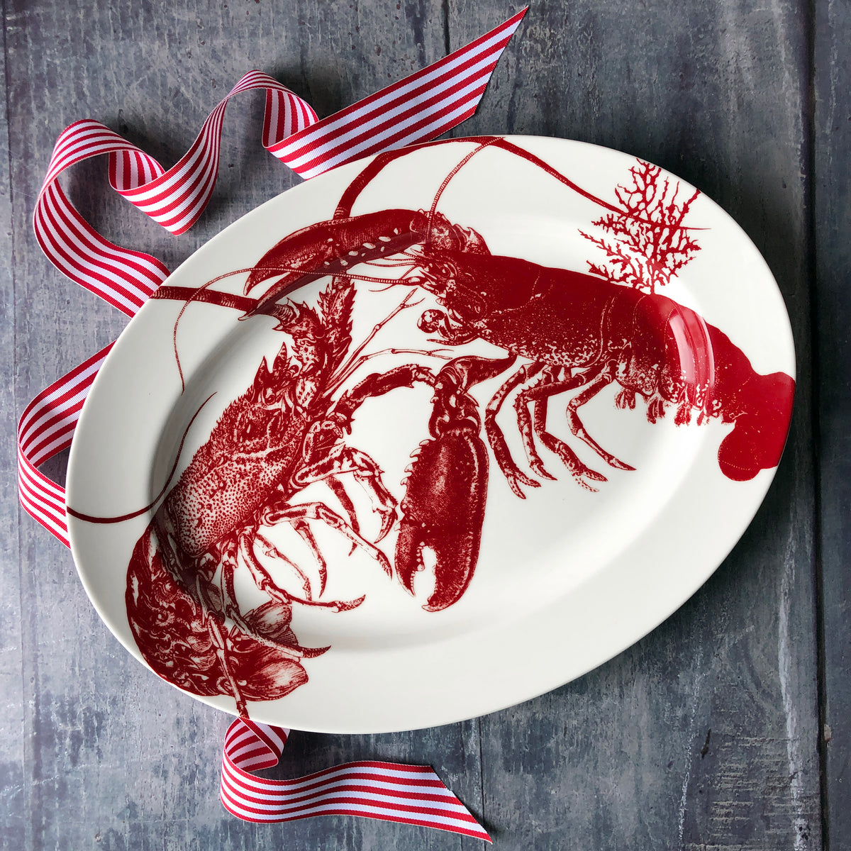 A white oval Father&#39;s Day Bundle with red lobster designs sits elegantly on a dark wooden surface. Red and white striped ribbons are arranged decoratively around the plate, enhancing the nautical theme by Caskata.