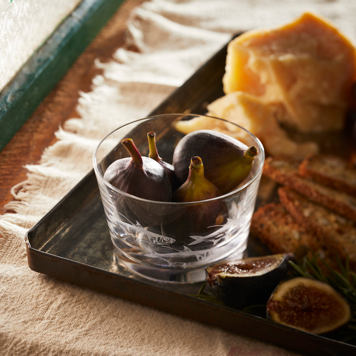 A clear glass bowl containing figs is on a metal tray with cheese, bread, and herbs, all placed on a beige cloth surface. This elegant setup perfectly complements the Caskata Lake House Bundle decor, adding a touch of rustic charm.